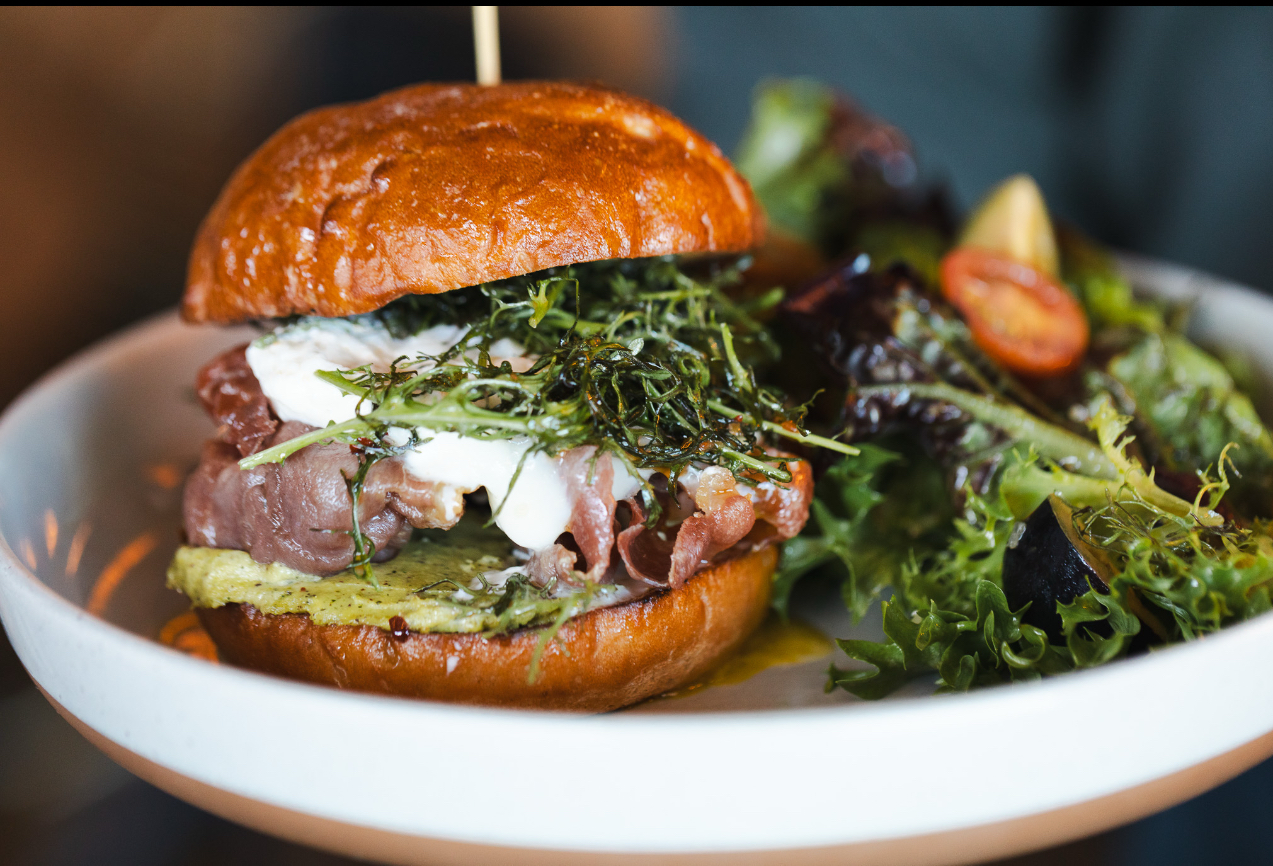 A finished brioche sandwich with prosciutto, burrata and crisp frisée salad served in a shallow bowl