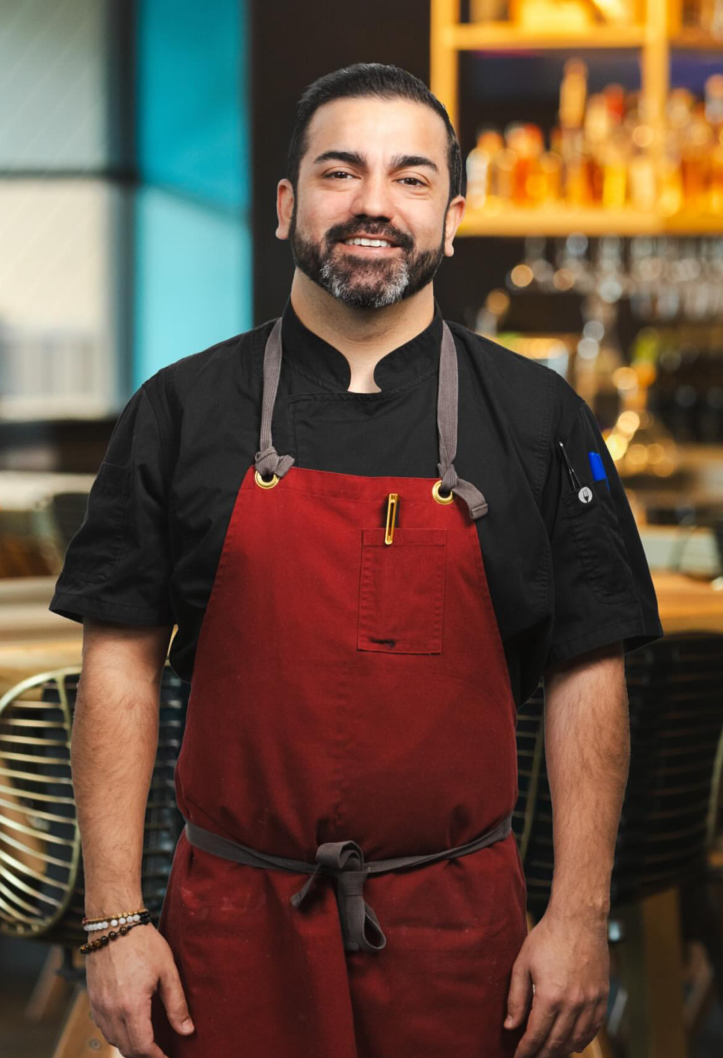 Chef Sebastian in a black chef coat and red apron, smiling in a warmly lit restaurant kitchen