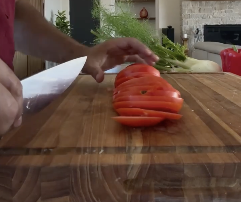 A chef's knife slicing ripe tomatoes on a walnut board with fresh fennel alongside