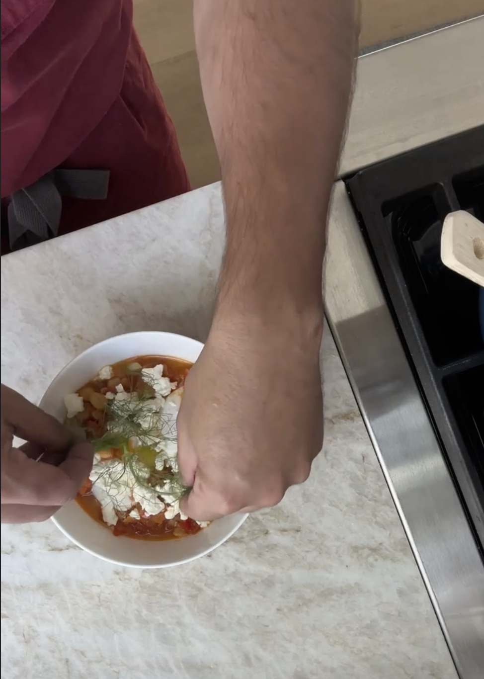 Hands finishing a bowl of stewed tomatoes, beans, feta and fennel fronds beside the range